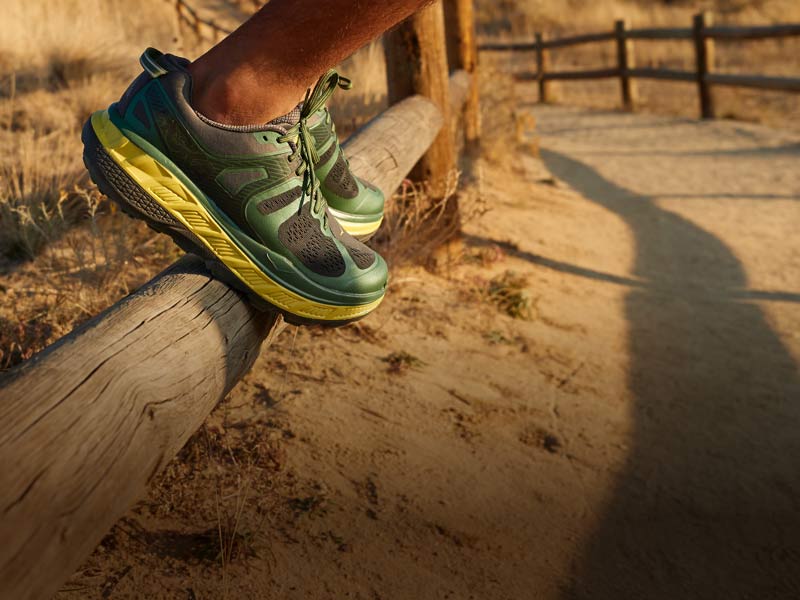 Person standing on wooden fence wearing HOKA shoes.